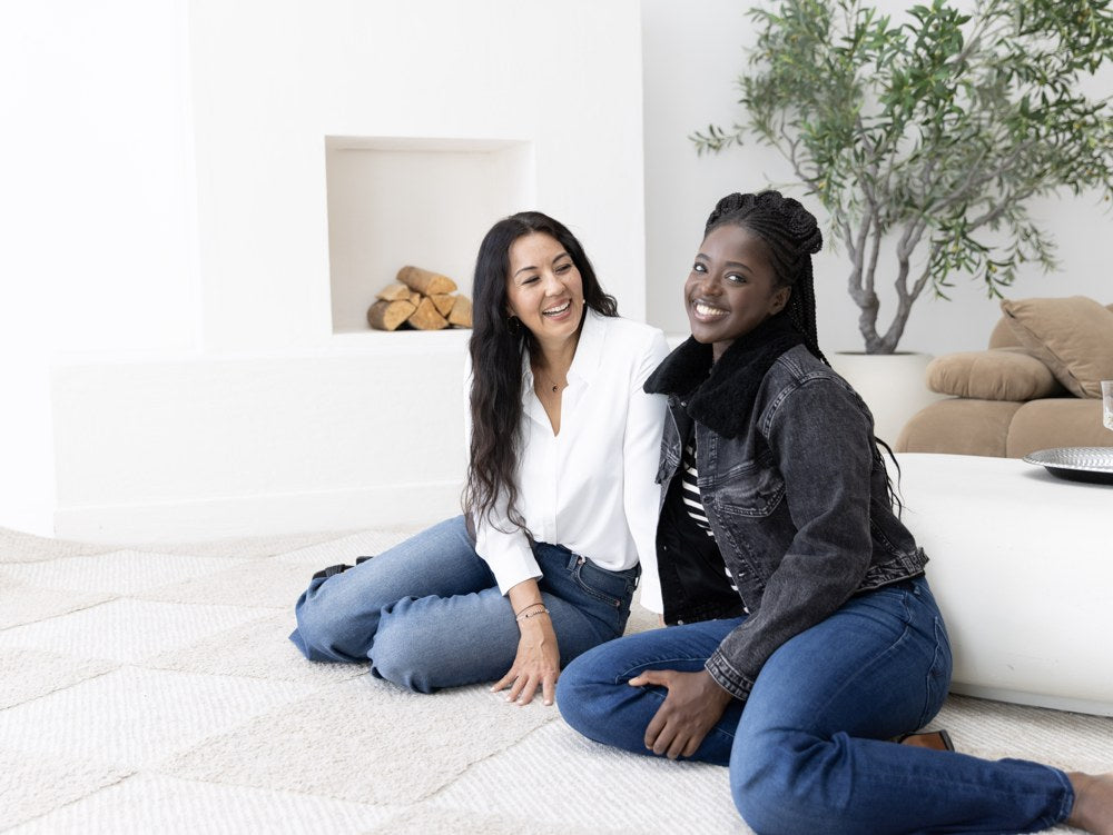 Two women laughing in relaxed denim outfits beside the sofa